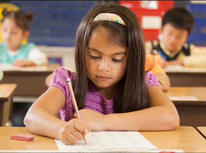 Young students at desks writing, centered on brunette girl.